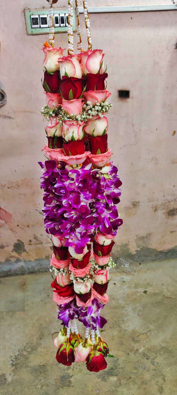 Garland of red and peach roses with a purple orchid section and red rose tassels, hanging against a wall.