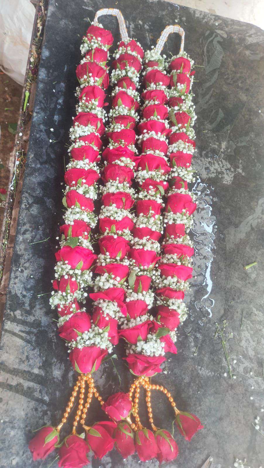 Two red rose and baby's breath garlands with white cord hangers and gold bead tassels of red roses, laid on stone.