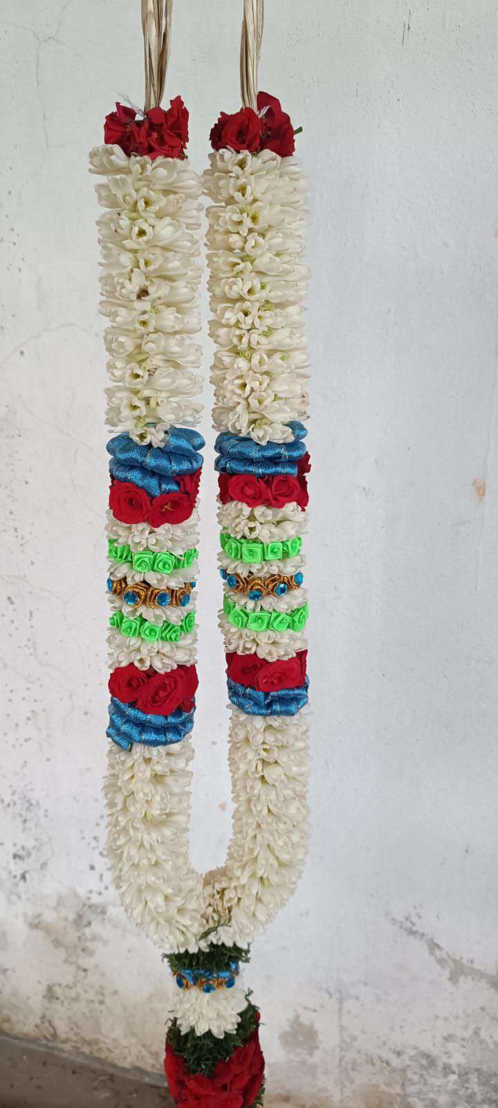 Tuberose garland with red, blue, green and gold bands and a red rose tassel, hanging against a wall.