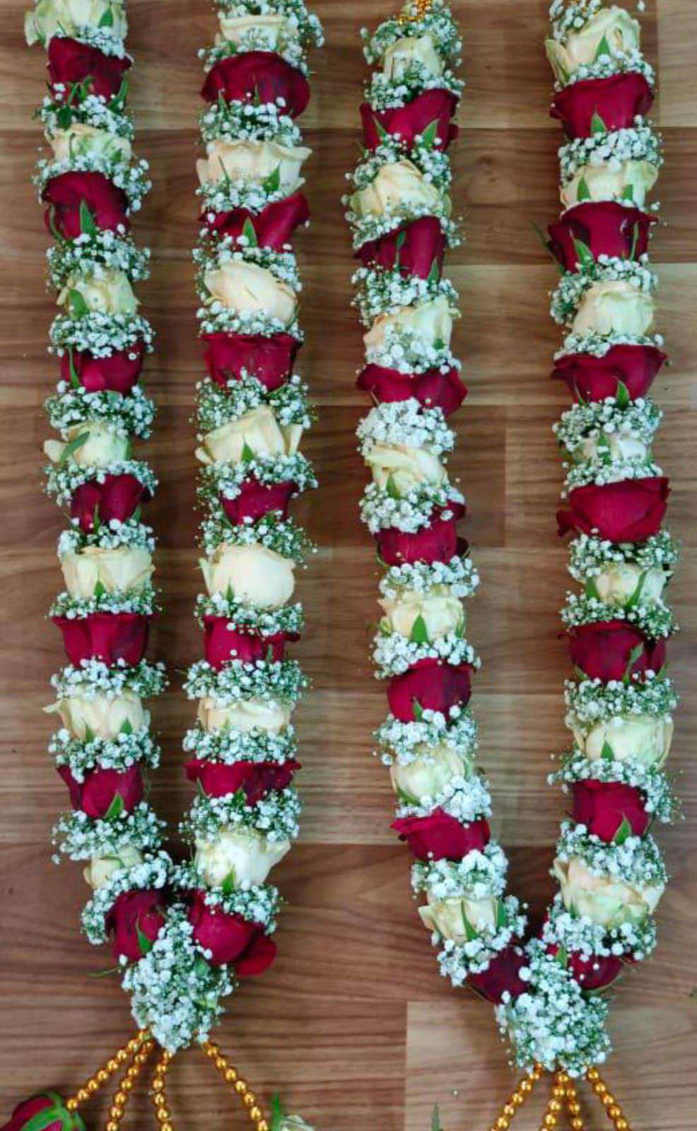 Two red and white rose garlands with baby's breath laid flat on wood, with gold bead tassels.