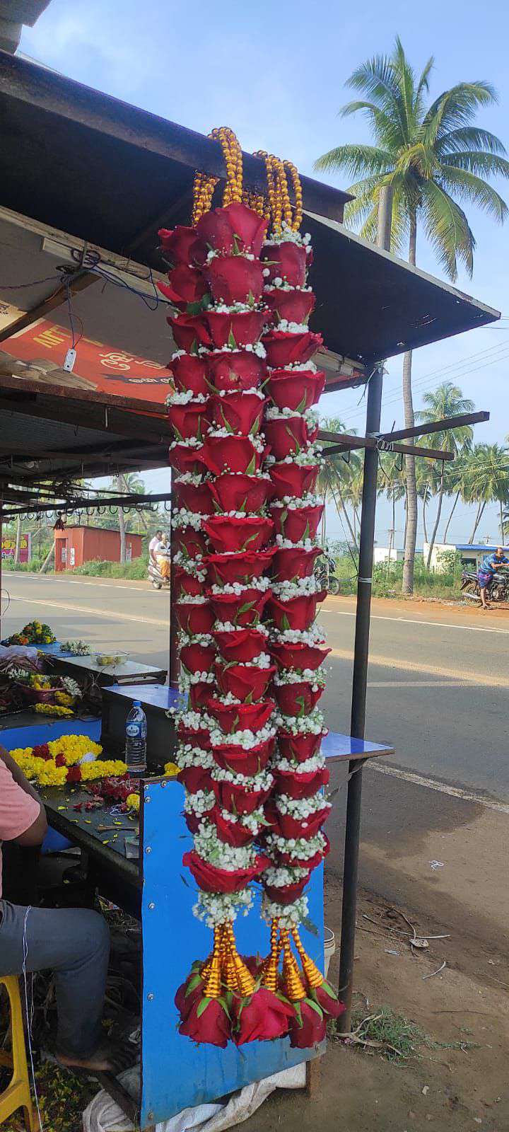 Pair of red rose and baby's breath garlands with gold bead hangers and gold-cone red rose tassels, hanging at a roadside stall.