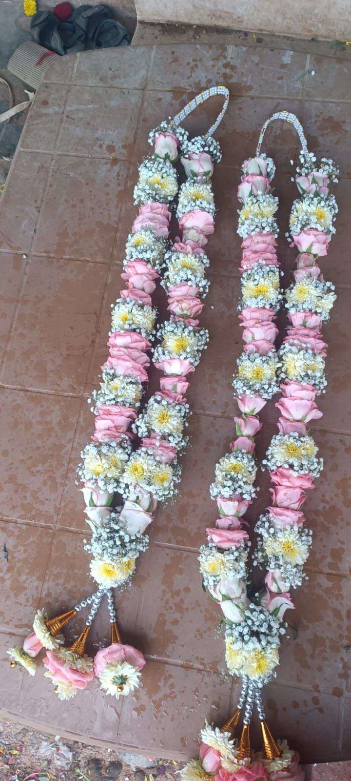 Two pink rose garlands with white daisy-like flowers and baby's breath, laid flat with gold-cone tassels.