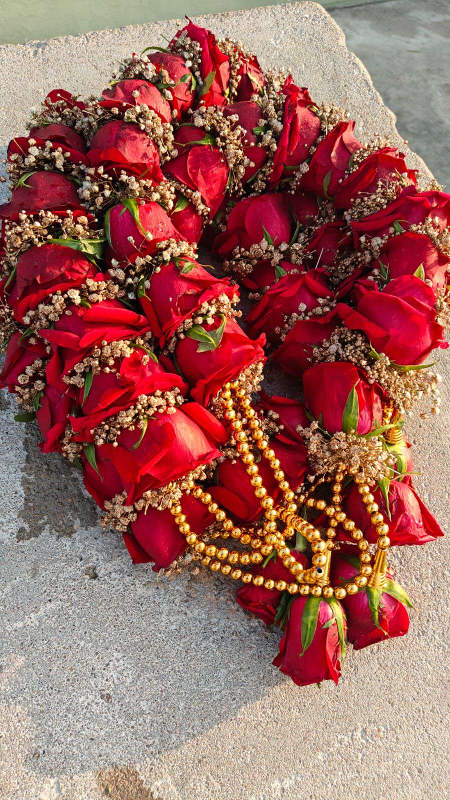 Red rose garland with baby's breath and long gold bead strands, laid in a coil on a stone surface.