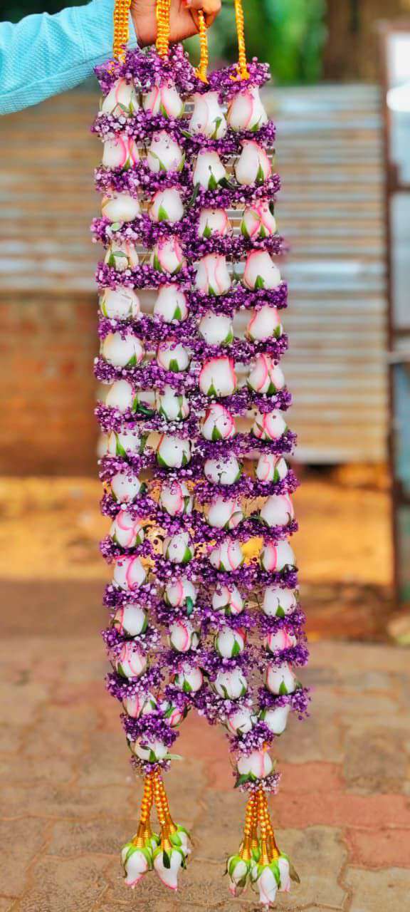 White rose-like blooms edged with pink, separated by small purple flowers, in hanging garlands with gold-coloured tassels.