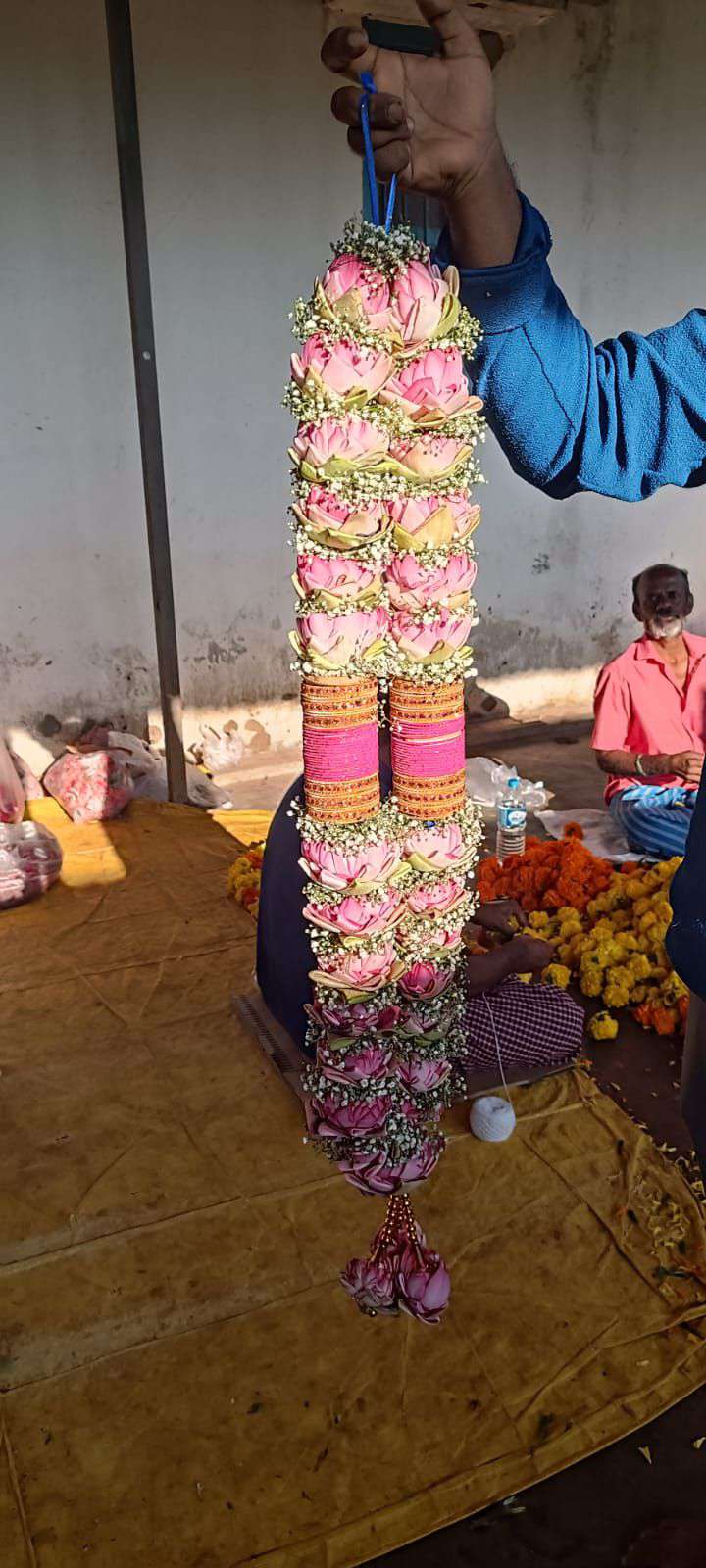 Pink lotus garland with a pink and gold band, held up in a flower market.